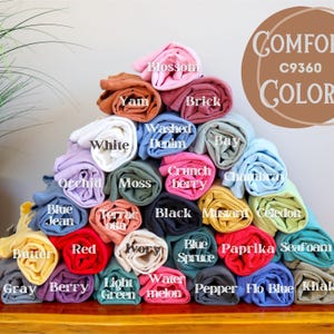 May include: A pyramid of rolled-up t-shirts in various colors, including white, blue, red, and yellow. Each shirt is labeled with its color name, such as "Blossom," "Yam," and "Washed Denim." The image also features the text "Comfort C9360 COLOR."