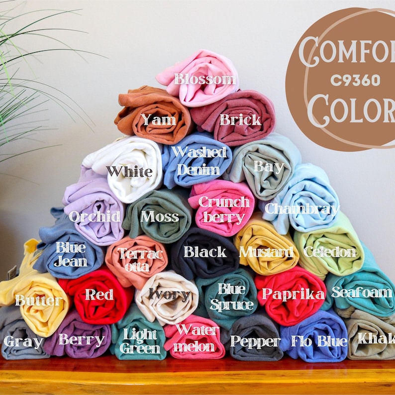 May include: A pyramid of rolled-up t-shirts in various colors, including white, blue, red, and yellow. Each shirt is labeled with its color name, such as "Blossom," "Yam," and "Washed Denim." The image also features the text "Comfort C9360 COLOR."