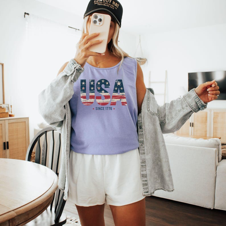 May include: A light purple tank top with the word "USA" in a patriotic design, featuring the American flag. The text "SINCE 1776" is below the word "USA". The person is wearing a black baseball cap and white shorts.