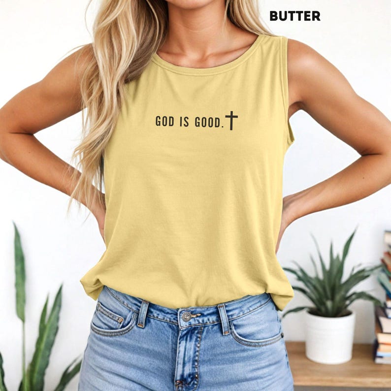 May include: A sleeveless, butter-colored tank top with the text "GOD IS GOOD." and a cross symbol in black. The top is paired with blue denim jeans. The word "BUTTER" is printed at the top of the image.