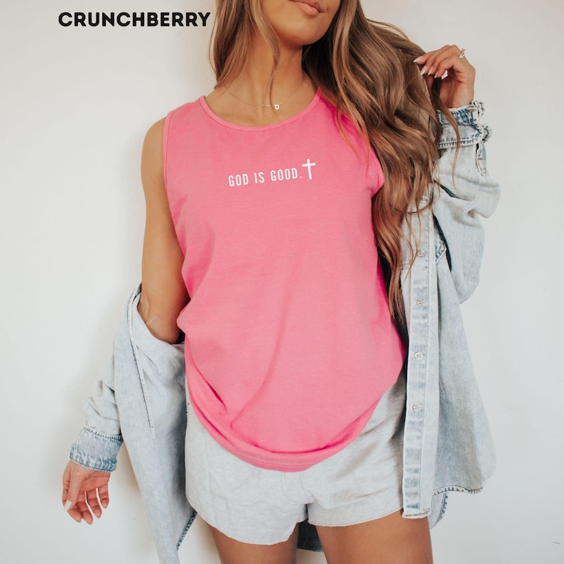 May include: A pink tank top with the text "GOD IS GOOD" and a cross graphic. The top is sleeveless and made of a soft material. The word "CRUNCHBERRY" is in the top left corner.