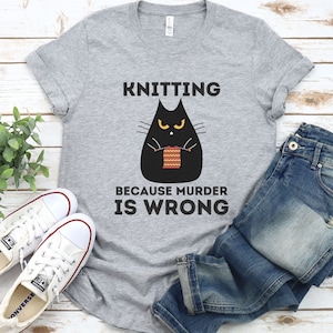 May include: Gray t-shirt with a black cat knitting a red and yellow scarf. The text on the shirt reads "Knitting because murder is wrong."
