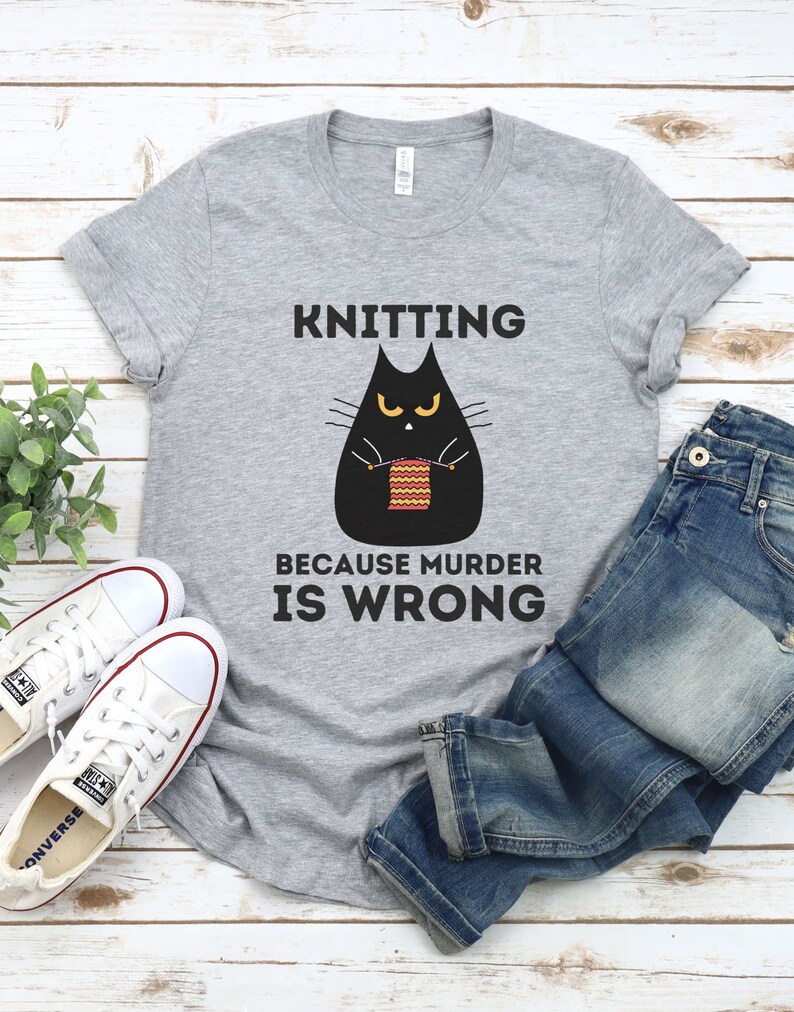 May include: Gray t-shirt with a black cat knitting a red and yellow scarf. The text on the shirt reads "Knitting because murder is wrong."