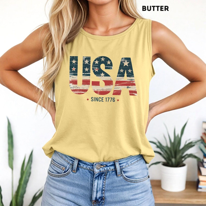 May include: A yellow tank top with the word "USA" in red, white, and blue, with a distressed American flag design. The text "SINCE 1776" is below the word "USA". The tank top is made of a soft material.