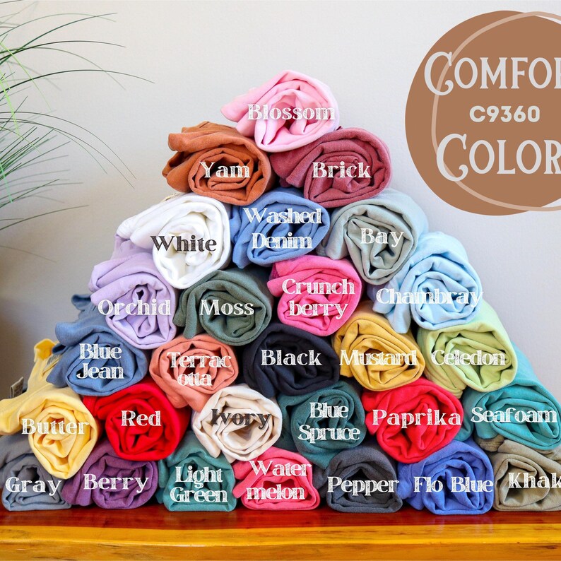 May include: A pyramid of rolled-up t-shirts in various colors, including white, blue, red, and yellow. Each shirt is labeled with its color name, such as "Blossom," "Yam," and "Washed Denim." The image also features the text "Comfort C9360 COLOR."