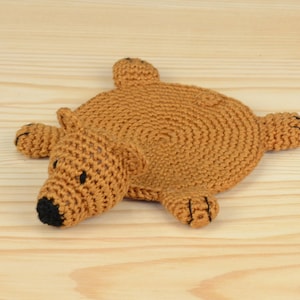 May include: A brown crocheted bear-shaped coaster with black eyes and nose. The coaster is lying on a light wood surface.