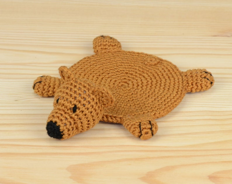 May include: A brown crocheted bear-shaped coaster with black eyes and nose. The coaster is lying on a light wood surface.