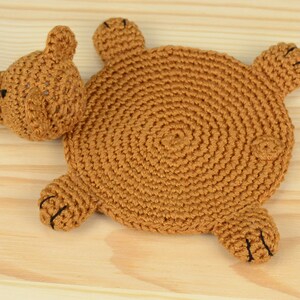 May include: A brown crocheted bear-shaped coaster with black eyes and nose. The coaster is lying on a light brown wooden surface.