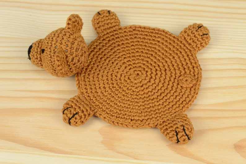 May include: A brown crocheted bear-shaped coaster with black eyes and nose. The coaster is lying on a light brown wooden surface.