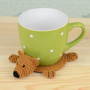 May include: A green mug with white polka dots sits on a crocheted brown bear coaster. The bear has a black nose and is lying on its belly.