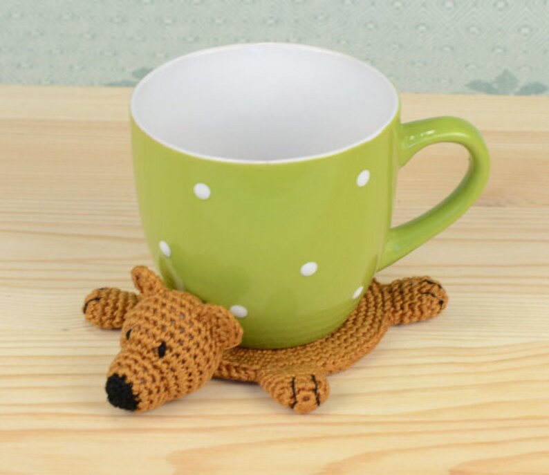 May include: A green mug with white polka dots sits on a crocheted brown bear coaster. The bear has a black nose and is lying on its belly.