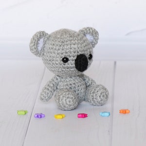 May include: A grey crocheted koala plush toy sitting on a white wooden surface with colourful star-shaped beads scattered around it.