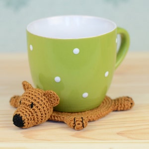 May include: A crocheted brown bear coaster with a green polka dot mug on top. The coaster is shaped like a bear with a brown nose and black eyes.