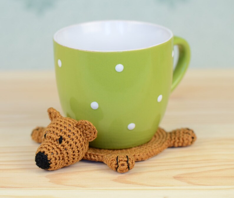 May include: A crocheted brown bear coaster with a green polka dot mug on top. The coaster is shaped like a bear with a brown nose and black eyes.