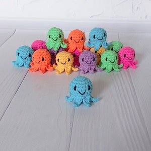 May include: A collection of colorful, crocheted octopus toys in various shades, including blue, orange, green, pink, yellow, and purple. Each octopus has a smiling face and is arranged on a white surface.