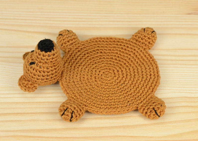 May include: A brown crocheted bear-shaped coaster with black eyes. The coaster is lying on a light brown wooden surface.