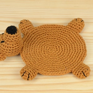 May include: A brown crocheted bear-shaped coaster with black eyes and a brown nose. The coaster is lying on a light brown wooden surface.