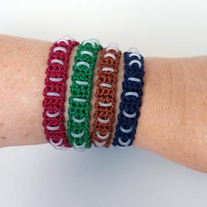 May include: Four woven bracelets in burgundy, green, brown, and navy blue. Each bracelet features a series of small, translucent white plastic rings integrated into the centre of the corded design, creating a repeating pattern along the length.