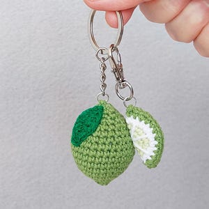 Può includere: Un portachiavi a forma di lime fatto a mano all'uncinetto, con un corpo verde e un accento di foglia verde. Una fetta di lime è attaccata al portachiavi. Il portachiavi è attaccato a un portachiavi argentato.