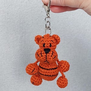 May include: Crocheted orange tiger keychain with black stripes and a black nose. The handmade tiger has a silver-colored clasp for attaching to keys or bags. The tiger's arms and legs are small orange balls.