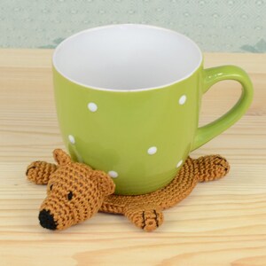 May include: A green mug with white polka dots sits on a crocheted brown bear coaster. The bear has a brown nose and black eyes.