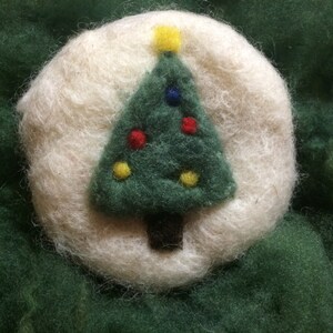 Op de afbeelding: Een witte vilten ornament met een groene vilten kerstboom met gele en rode vilten ornamenten.