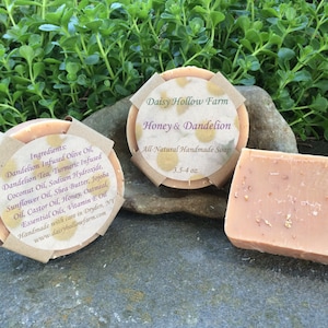 May include: Two bars of handmade soap, one round and one rectangular, with a label that reads "Daisy Hollow Farm Honey & Dandelion All-Natural Handmade Soap 3.5-4 oz". The soap is a light peach color and has a natural, rustic look.