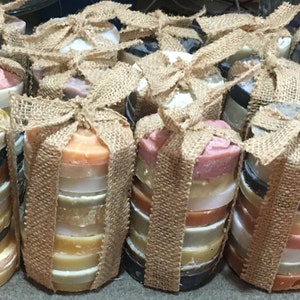 Puede incluir: Una colección de jabones redondos multicolores envueltos en arpillera y atados con cuerda. Los jabones están apilados en columnas y dispuestos en una fila.