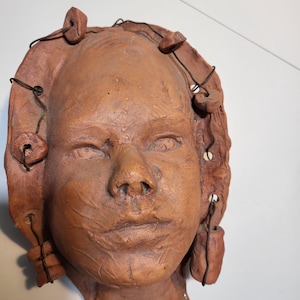 May include: A terracotta sculpture of a human face with detailed features. The face has a textured surface and is adorned with wire and small, rounded clay elements. The sculpture is a warm, earthy tone and appears to be a wall hanging.