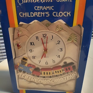 May include: A ceramic children's clock with a white clock face and red hands. The clock features a train design with the words "ALRINE CHOO CHOOTRAIN". The clock is in a blue box with the words "Sunbeam Quartz Ceramic Children's Clock".