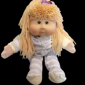 May include: A Cabbage Patch Kid doll with blonde yarn hair and a purple butterfly hair clip. The doll is wearing a gray and white striped sleeper.