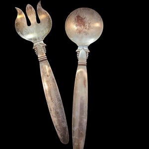 May include: A set of two silver-plated serving utensils, a fork and a spoon, with ornate handles. The utensils have a vintage look and show signs of wear.