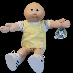 May include: A Cabbage Patch Kids doll with a bald head and blue eyes. The doll wears a yellow corduroy overall and a white shirt. The doll has white sneakers with black stripes and a hang tag.