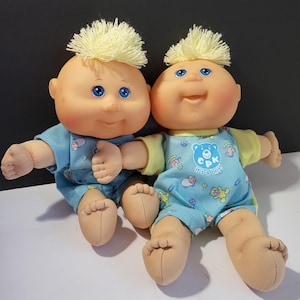 May include: Two Cabbage Patch Kids dolls. Each doll has a bald head with yellow yarn hair, blue eyes, and a smiling expression. They wear blue and yellow onesies with a pattern of sheep and the text "It's a Boy!"