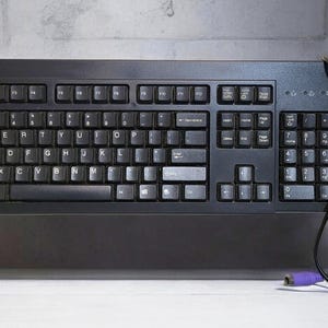 May include: A black IBM keyboard with a full layout, including a numeric keypad. The keyboard has a wired connection with a purple connector. The keys are labeled with white letters and symbols. The IBM logo is visible on the top left.
