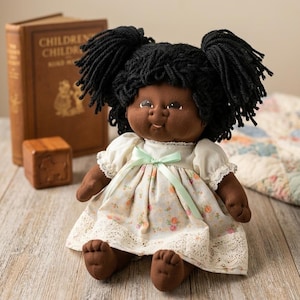 May include: A handmade doll with dark skin, black yarn hair in two high pigtails, and brown fabric arms and legs. The doll wears a floral print dress with a lace hem and a mint green bow. A vintage book is in the background.