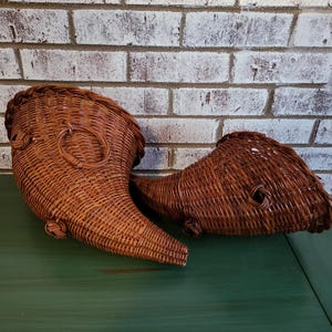 May include: Two brown woven wicker cornucopia baskets with a curved horn shape. Each basket features a wide, open top and a tapered, pointed end, with a circular woven ring attached to the side of the larger basket for hanging or display.