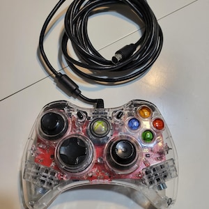 May include: Clear, wired Xbox 360 controller with a black cable. The controller has a transparent shell, revealing the internal red circuit board and colored buttons. The controller is labeled "AFTERGLOW".