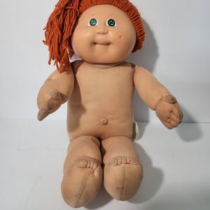 May include: A vintage Cabbage Patch Kid doll with a peach-colored face, green eyes, and orange yarn hair. The doll is nude, with a soft body and fabric limbs. The doll is sitting down.