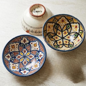 May include: Three ceramic bowls with intricate floral patterns. One bowl is white with black and red accents, while the other two feature vibrant designs in blue, yellow, and brown. The bowls have a rustic, handcrafted appearance.
