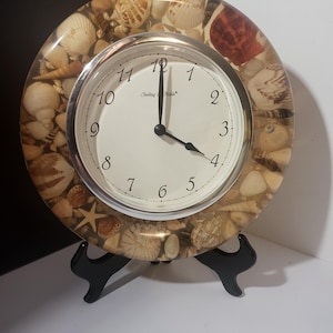 May include: A round clock with a white face and black hands, set on a black stand. The clock's bezel is filled with various seashells. The clock face has black numerals and the brand name "Sterling & Noble".