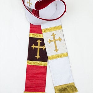 Könnte beinhalten: Eine weiße und rote liturgische Stola mit goldenen Verzierungen und gestickten Kreuzen. Die Stola hat unten einen Fransen.