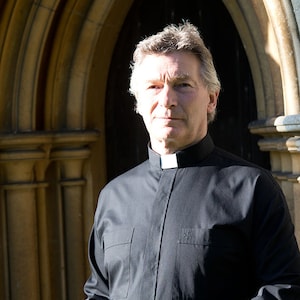 May include: A man wearing a black clerical shirt with a white collar stands in front of a stone archway. He is holding a closed book in his hands.