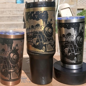 May include: Three insulated travel mugs with a train design. The mugs are in varying sizes and colors, including copper, black, and gold. Each mug features a detailed engraving of a vintage steam train, with smoke billowing from the smokestack.