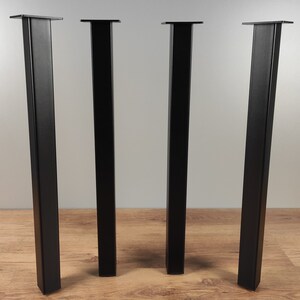 May include: Four black metal table legs with square tops. The legs are standing on a wood floor.