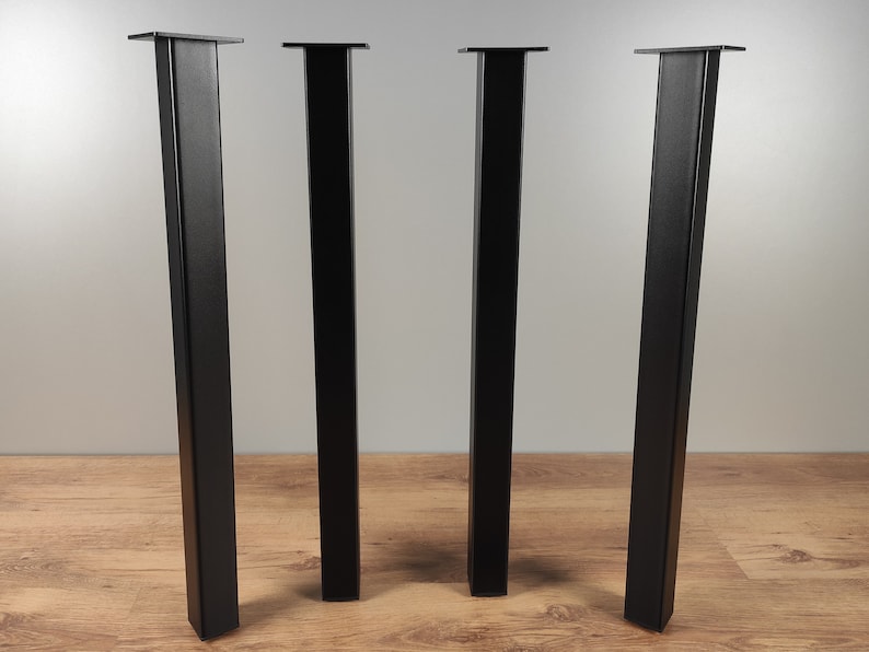 May include: Four black metal table legs with square tops. The legs are standing on a wood floor.