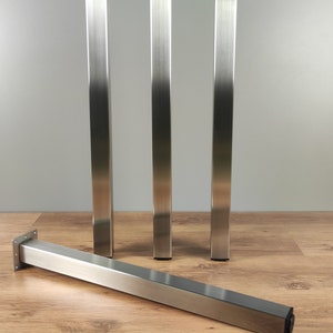 May include: Four brushed stainless steel table legs. Three legs are standing upright, and one leg is lying flat on a wood floor. The legs are square in shape and have a brushed finish.
