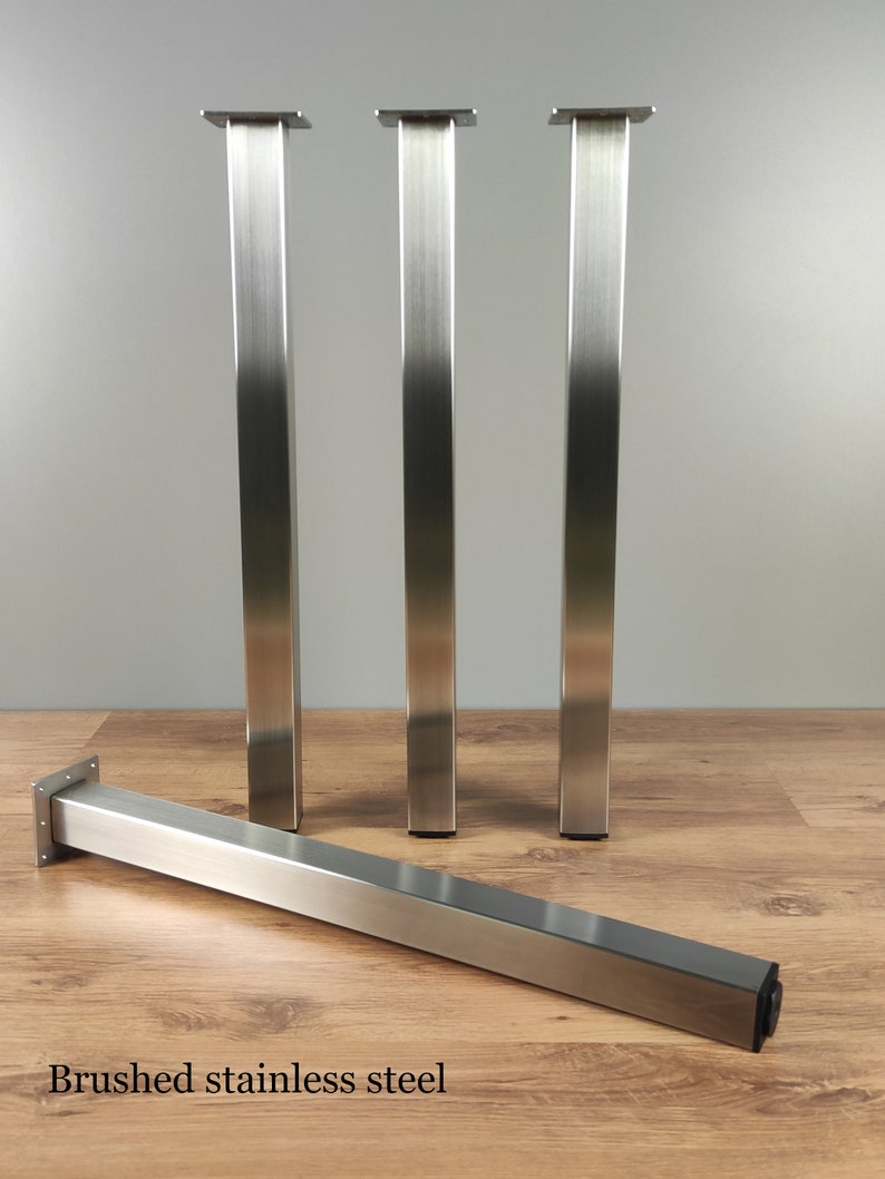 May include: Four brushed stainless steel table legs. Three legs are standing upright, and one leg is lying flat on a wood floor. The legs are square in shape and have a brushed finish.