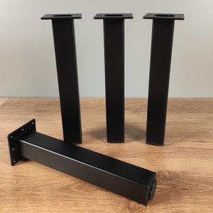 May include: Four black metal table legs with a square top and a rectangular base. The legs are standing on a wooden surface.