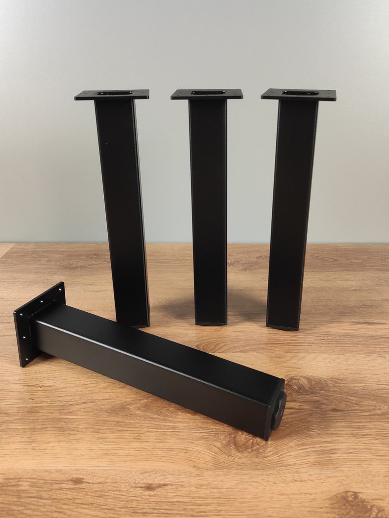 May include: Four black metal table legs with a square top and a rectangular base. The legs are standing on a wooden surface.